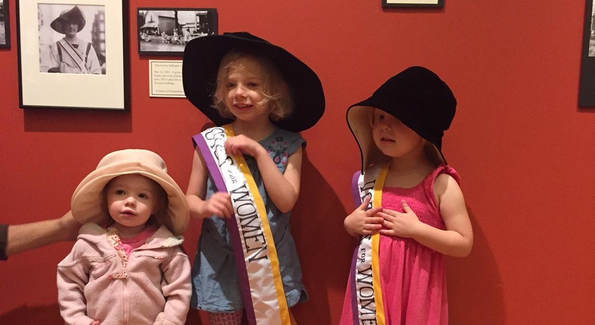 Children wear suffragette hats and sashes.