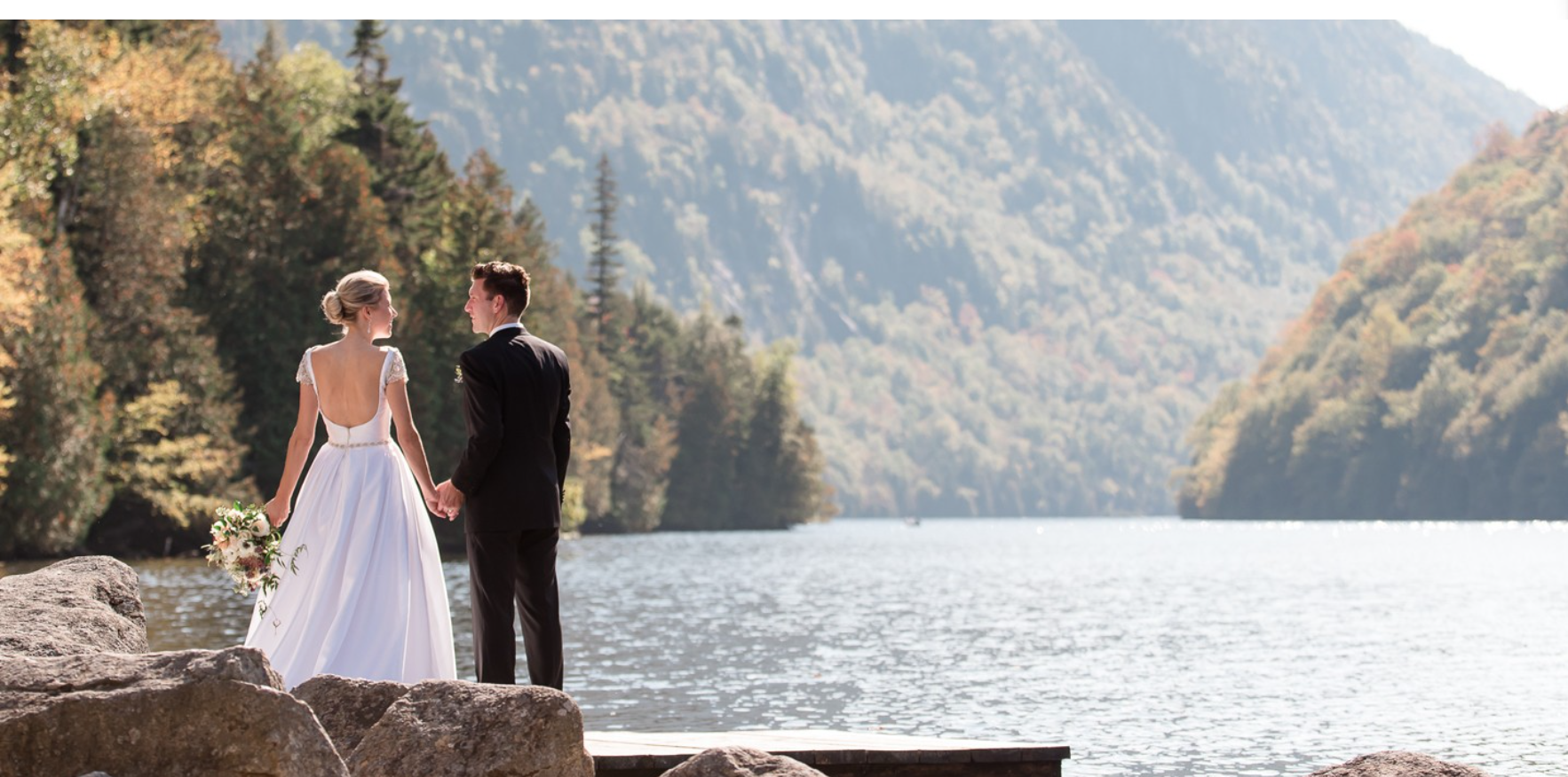 A bride and groom stand on a dock by the water surrounded by pine covered mountains.