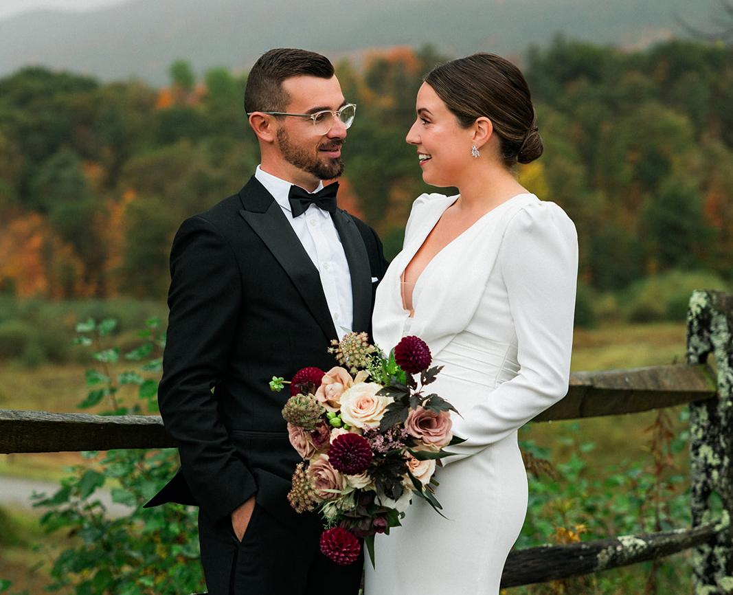 Bride and groom stand near a fence while she holds a bouquet of deep colored roses.