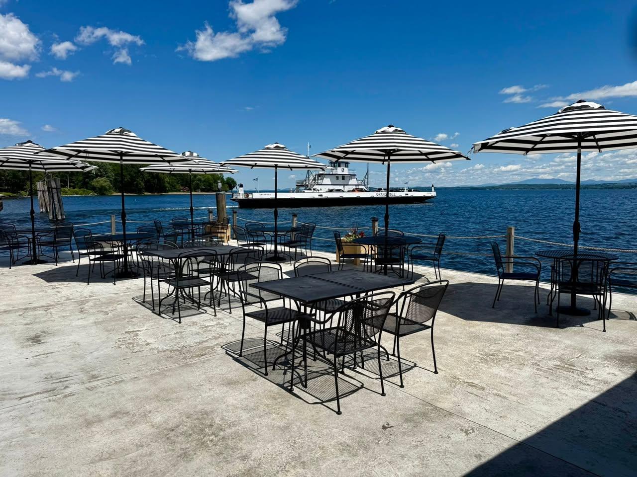 Metal tables and chairs with black and white striped sun umbrellas on the deck looking over Lake Champlain.